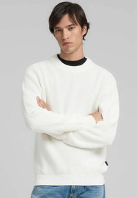 Maglione bianco in maglia testurizzata con scollo rotondo e polsini a costine. Indossato sopra una camicia nera, presenta una discreta targhetta con logo sul fondo.