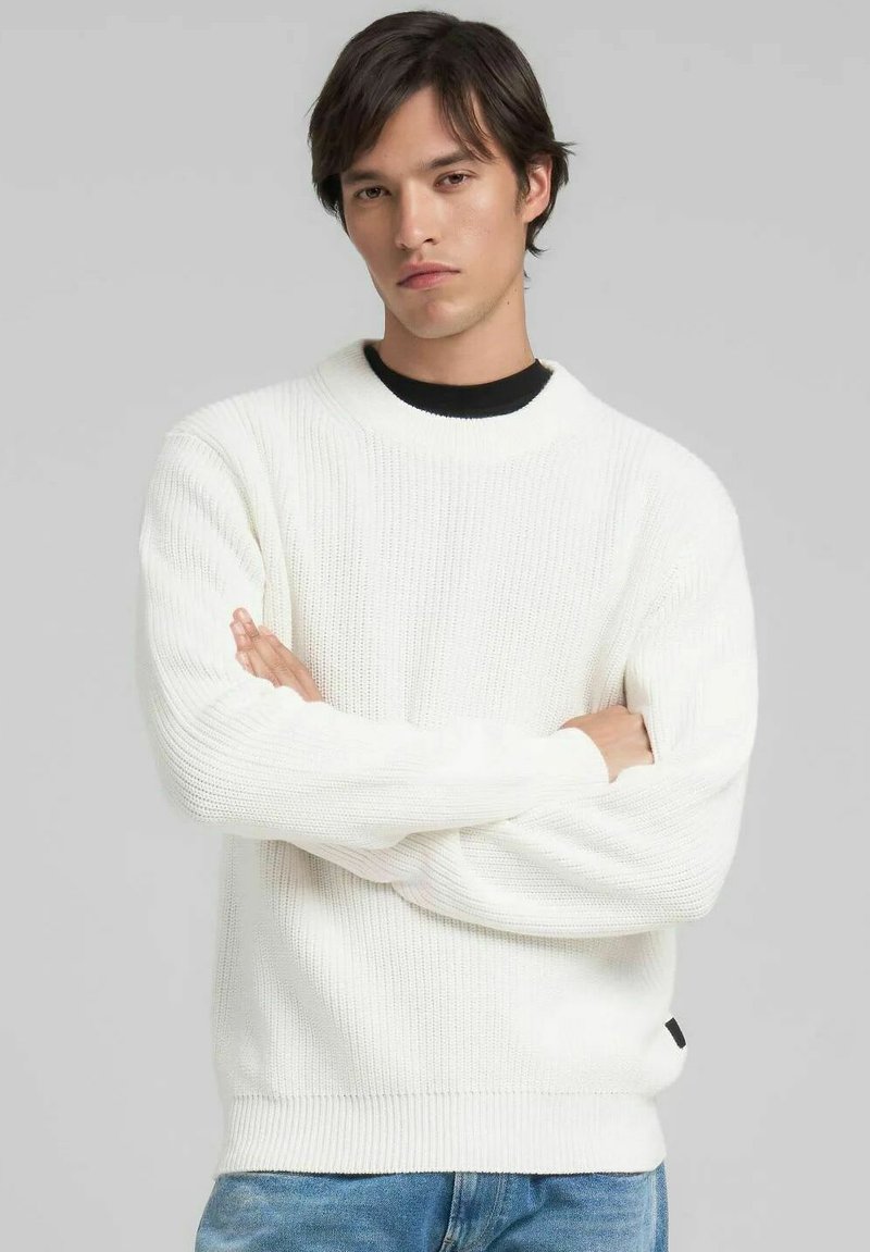 Maglione bianco in maglia testurizzata con scollo rotondo e polsini a costine. Indossato sopra una camicia nera, presenta una discreta targhetta con logo sul fondo.