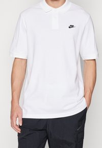 Valkoinen polo paita, joka on valmistettu puuvillasta, klassisella kauluksella, lyhyillä hihoilla ja pienellä mustalla Nike-logolla vasemmassa rinnassa.