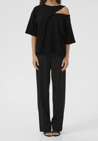 Sort oversized kortærmet top med asymmetrisk design, båret med skræddersyede sorte bukser og åbne sandaler.