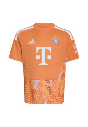 Niño sonriente que lleva puesta la camiseta y pantalones cortos de fútbol naranja del FC Bayern Múnich con patrones abstractos blancos y morados, de pie frente a un fondo liso.