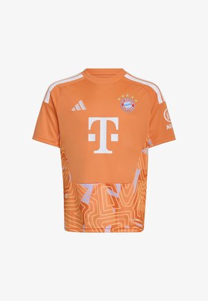 Niño sonriente que lleva puesta la camiseta y pantalones cortos de fútbol naranja del FC Bayern Múnich con patrones abstractos blancos y morados, de pie frente a un fondo liso.