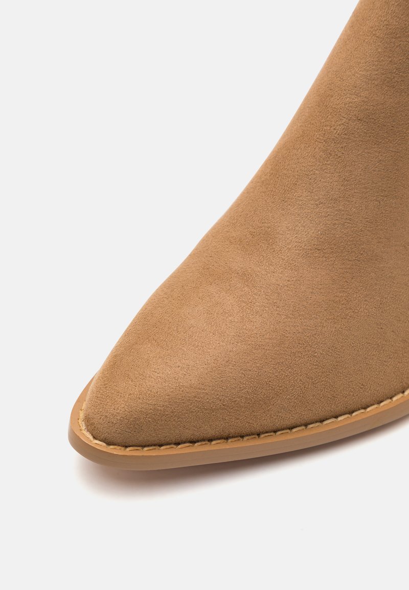 Beżowe zamszowe botki za kostkę z wydłużonym noskiem, subtelnymi detalami przeszyć oraz solidną beżową gumową podeszwą. Gładka tekstura na całej powierzchni.