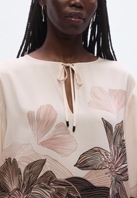 Blouse rose clair avec imprimé floral dans des tons doux ; un col en trou de serrure avec un détail de lien et des accents foncés contrastants. Texture lisse.