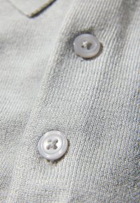 Jasnoszary dzianinowy materiał o fakturowanej powierzchni, z dwoma jasnymi guzikami w czterdziurkowym wzorze. Zbliżenie na obszar kołnierza.