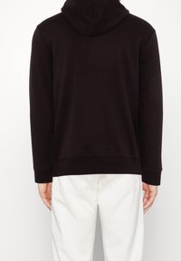 Felpa nera con cappuccio e texture uniforme, caratterizzata da orlo e polsini a costine, vista da dietro, abbinata a pantaloni chiari.