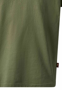 Section de tissu vert olive d'un t-shirt avec une petite étiquette latérale rouge foncé visible sur le côté droit.