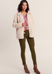 Giacca in pile color beige chiaro con colletto, chiusura a bottoni e tasche laterali. Indossata sopra una maglietta floreale colorata e pantaloni verde oliva. Stivaletti marroni alla caviglia.