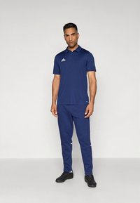 Marineblaues Poloshirt mit Kragen und Adidas-Logo, kombiniert mit passenden marineblauen Sporthosen mit weißen Akzenten. Dazu werden schwarze Sportschuhe getragen.