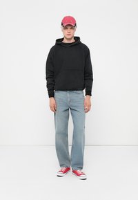 Svart fleece hettegenser med frontlomme, lyseblå rettbente jeans og røde kanvas sneakers, kombinert med en rosa baseballcaps.