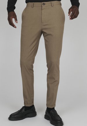 Pantalon beige ajusté avec passants de ceinture et bouton à l'avant, porté avec des chaussettes noires et des mocassins en cuir noir sur une personne à la peau foncée.
