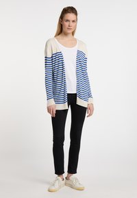 Cardigan a righe blu e crema, con design aperto sul davanti, maniche lunghe, realizzato in un tessuto leggero, abbinato a pantaloni skinny neri e sneakers bianche.