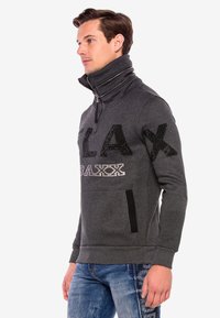 Grau gepolsterter Hoodie mit hohem Kragen, ausgestattet mit schwarzem Pailletten-Text "PLAX" und "BAXX" sowie einer Seitentasche mit schwarzem Akzent.