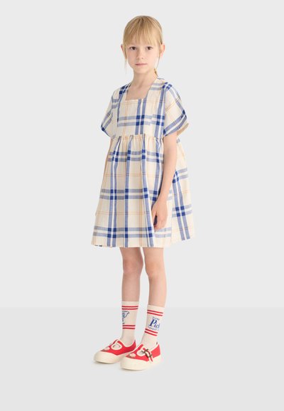 Jeune fille portant une robe à manches courtes, longueur genou, à carreaux bleus et beiges, avec des chaussettes blanches ornées de rayures rouges et des chaussures rouges à boucles.