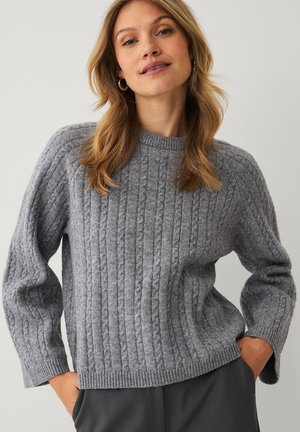 Kobieta o blond włosach, nosząca kolczyki koła i szary sweter warkoczowy, z rękami w kieszeniach ciemnych spodni.