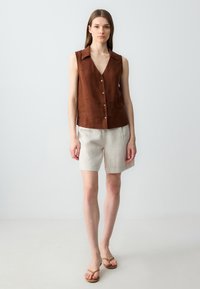 Top in lino marrone senza maniche con scollatura a V e bottoni, abbinato a pantaloni corti in lino beige chiaro. Il modello indossa semplici sandali marroni.