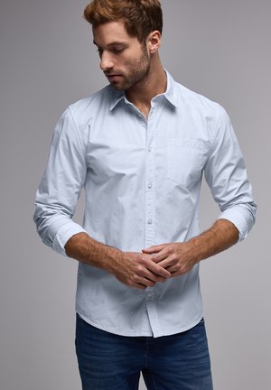 Lyseblå skjorte med knapper og et subtilt struktureret mønster, lange ærmer rullet op til manchetterne, og en lille brystlomme, båret med mørke jeans.