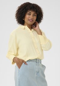 Camicia button-up gialla chiara a maniche lunghe con colletto e polsini arricciati, abbinata a una gonna di jeans. Texture morbida, vestibilità rilassata.