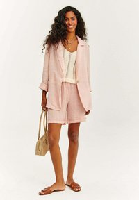 Světle růžový lněný blazer a odpovídající šortky s ohrnutými manžetami, doplněné krémovým pleteným topem a hnědými sandály na pásky, držící béžovou tašku.