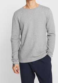 Chemise grise à manches longues avec texture côtelée, col rond et manches ajustées. Associée à un pantalon foncé. Étiquette de marque discrète à l'ourlet.