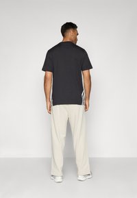 Homem vestindo uma t-shirt preta com riscas brancas nas laterais, calças cor creme e ténis brancos, de pé de frente para uma parede cinza clara lisa.