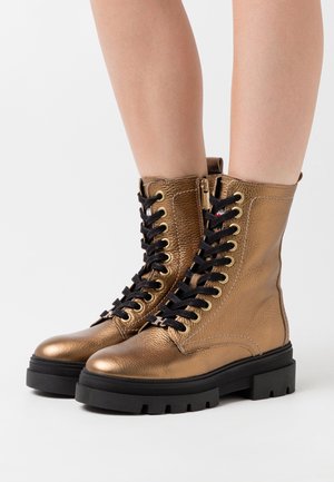 Pronssinväriset metallinhohtoiset nilkkurit, joissa on nyöritys, teksturoitu pinta, mustat nauhat, kultaiset reiät ja paksu musta kumipohja.