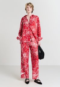 Jeune femme debout, vêtue d'une chemise et d'un pantalon à motifs floraux rouges et blancs, tenant un petit sac noir et portant des chaussures à talons noirs.