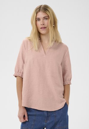 Blondynka w jasnoróżowej bluzce z krótkim rękawem i marszczonymi mankietami oraz niebieskich dżinsach, stojąca na białym tle.