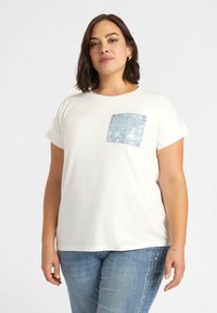 Camiseta blanca de manga corta con un bolsillo de lentejuelas azules texturizadas en el lado izquierdo del pecho, combinada con jeans de un azul claro. Tejido suave y corte relajado.