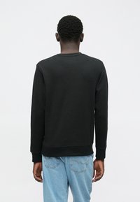 Osoba s krátkymi kučeravými vlasmi, oblečená v čiernej dlhoprstovej mikine a svetlomodrých džínsoch, stojí chrbtom k jednolitému pozadiu.