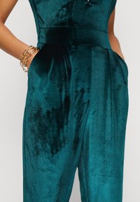 Teal sammetsoverall med en mjuk textur, med en figurnära midja, sidofickor och en subtil lyster. Guldkedjearmbandet framhäver looken.