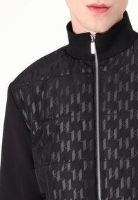 Svart pufferväst med hög krage, med ett geometriskt mönster och silverdragkedja. Slät textur med kontrasterande ärmmaterial.