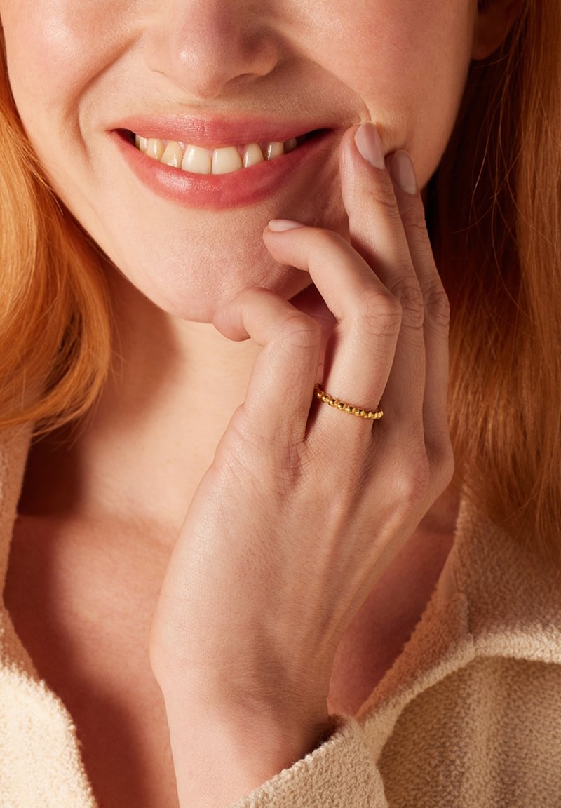 Gouden ring met een gestructureerd kraalontwerp, gedragen op een hand, met gladde huid en een neutraal gekleurd kledingstuk op de achtergrond.