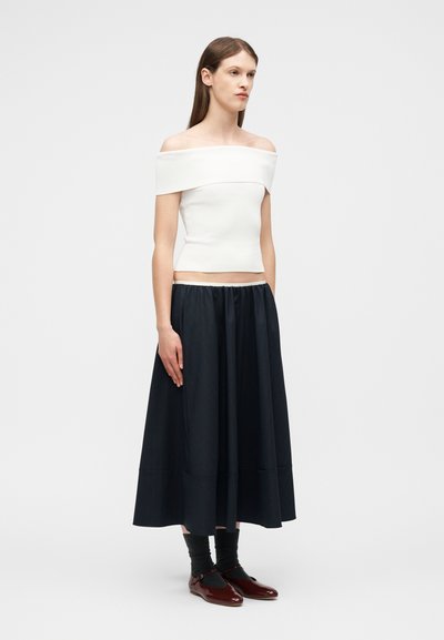 Femme aux longs cheveux bruns portant un haut blanc épaules dénudées, une jupe midi noire, des chaussettes noires et des chaussures Mary Jane rouge foncé, debout devant un fond uni.