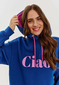 Modrá mikina s kapucí s nápisem "Ciao" v růžové barvě, která má růžovou podšívku kapuce a šňůrky. Hladký materiál, volný střih a dlouhé rukávy.