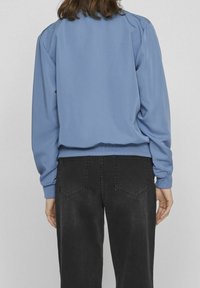 Blue sweatshirt with ribbed cuffs and hem, featuring a smooth texture. Paired with black jeans with two back pockets, showing a casual outfit.