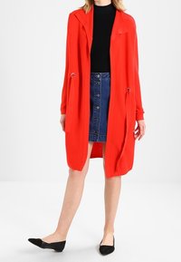 Cardigan rosso lavorato a maglia con collo sciallato, apertura frontale e vita regolabile, indossato sopra una maglietta nera e una gonna di jeans blu. Ballerine nere.