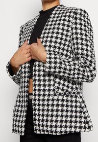 Svartvitt hounds-tooth-mönstrad blazer med en strukturerad design, kavajslag och två framfickor. Tyget ser texturerat ut.