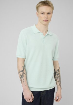 Jeune homme aux cheveux blonds courts portant un polo à manches courtes vert menthe clair et un jean foncé, montrant des tatouages floraux et de papillons sur les deux avant-bras.