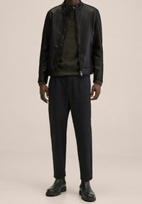 Zwarte leren jas met hoge kraag, groene textuurtrui en donkere broek, gecombineerd met zwarte enkellaarzen. Minimalistisch ontwerp.