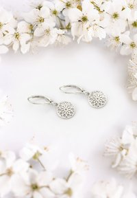 Silverhängande örhängen med cirkulär, utskuren blommönster. De hänger från hoop-spännen och har en slät, polerad yta.
