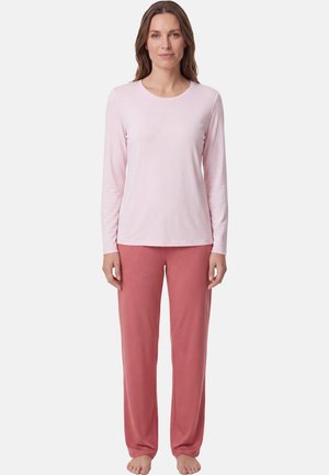 Vrouw staat blootsvoets en draagt een lichtroze top met lange mouwen en losse rozenkleurige broek tegen een effen witte achtergrond.