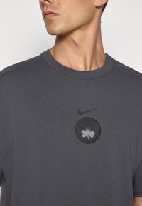 Mørkegrå t-shirt lavet af bomuld, med et rundt logo der har en kløver samt teksten "CELTICS." Indeholder et Nike-swoosh over logoet.
