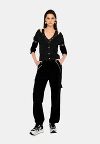 Cardigan nero con dettagli in pizzo, chiusura frontale con bottoni e maniche a tre quarti, abbinato a pantaloni neri con piping bianco, indossato con sneaker a fantasia.