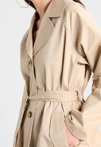 Donna che indossa un trench beige con cintura, colletto ampio, bottoni tartarugati e una mano in tasca, su sfondo bianco.