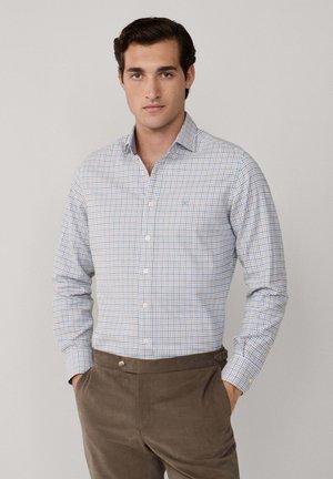 Camisa de botones en un patrón de cuadros azul y blanco, con cuello puntiagudo y mangas largas. Llevada con pantalones de pana marrones.