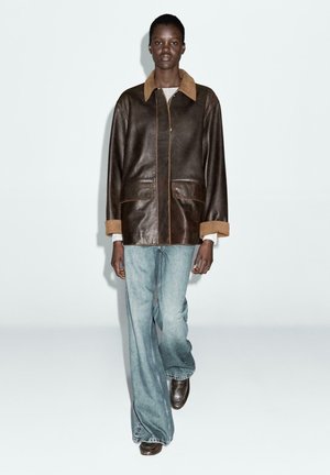 Brązowa skórzana kurtka z jasnobrązowym zamszowym kołnierzem, przednimi kieszeniami i kontrastowym przeszyciem, noszona na jasnoniebieskich dzwonach i brązowych butach.