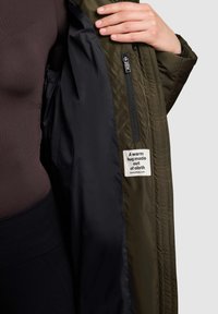 Green puffer jacket with black lining, visible zipper, and a fabric label stating "A warm hug made out of cloth." Smooth texture.