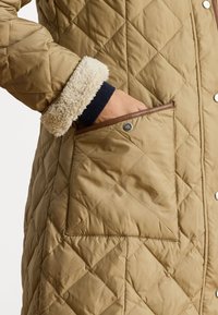 Klāts brūngans mētis ar mākslīgās kažokādas aproci, sānu kabatu ar brūnu apmali un pogu aizdari. Audumam ir gluda tekstūra.