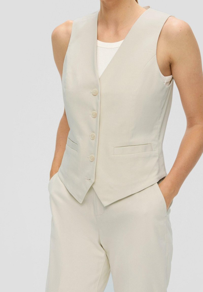 Femme portant un gilet beige sans manches boutonné sur un débardeur blanc, associé à un pantalon beige assorti, les mains dans les poches.